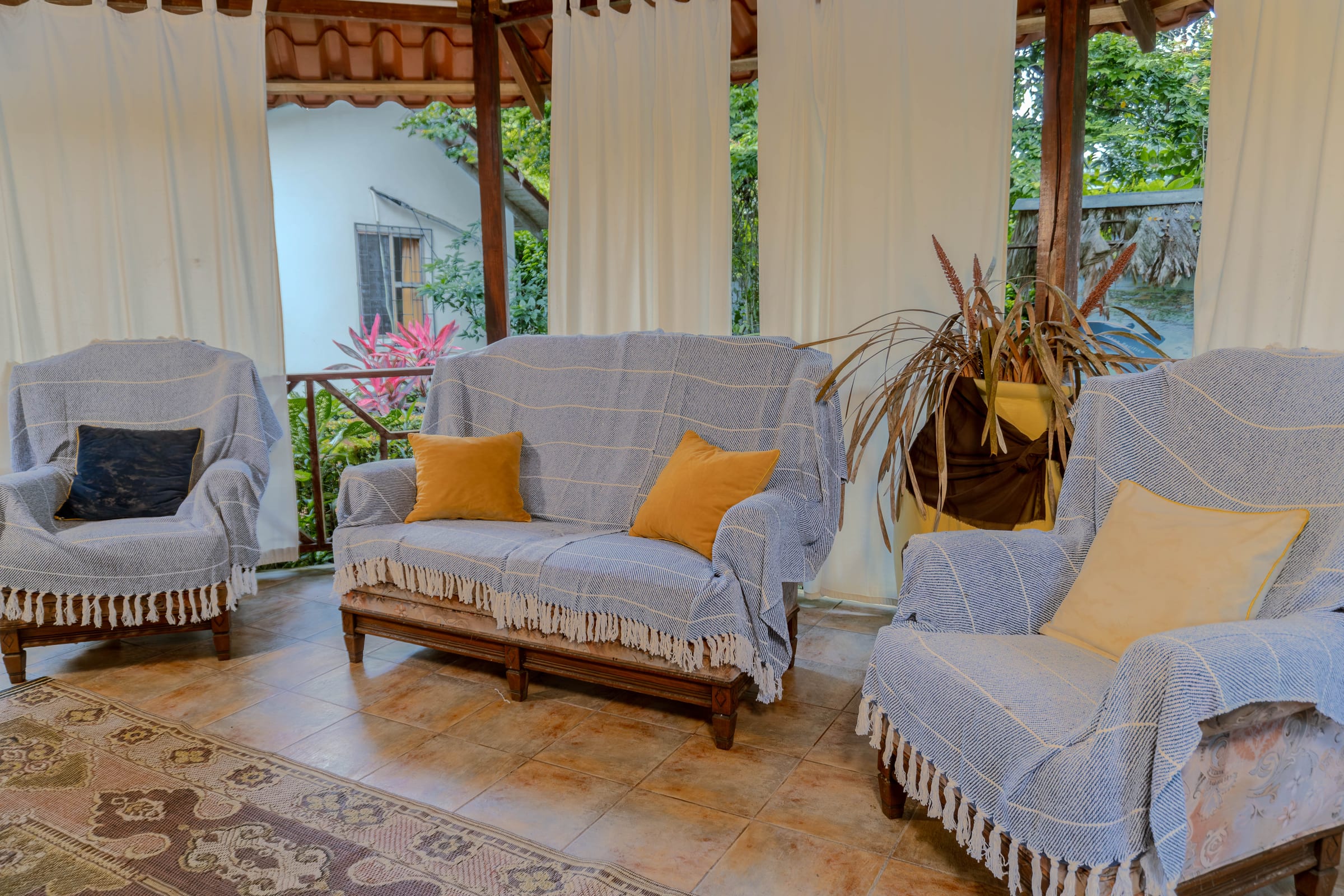 Covered lobby lounge with slipcovered sofas and armchairs, mustard-colored throw pillows, and sheer curtains framing a garden view