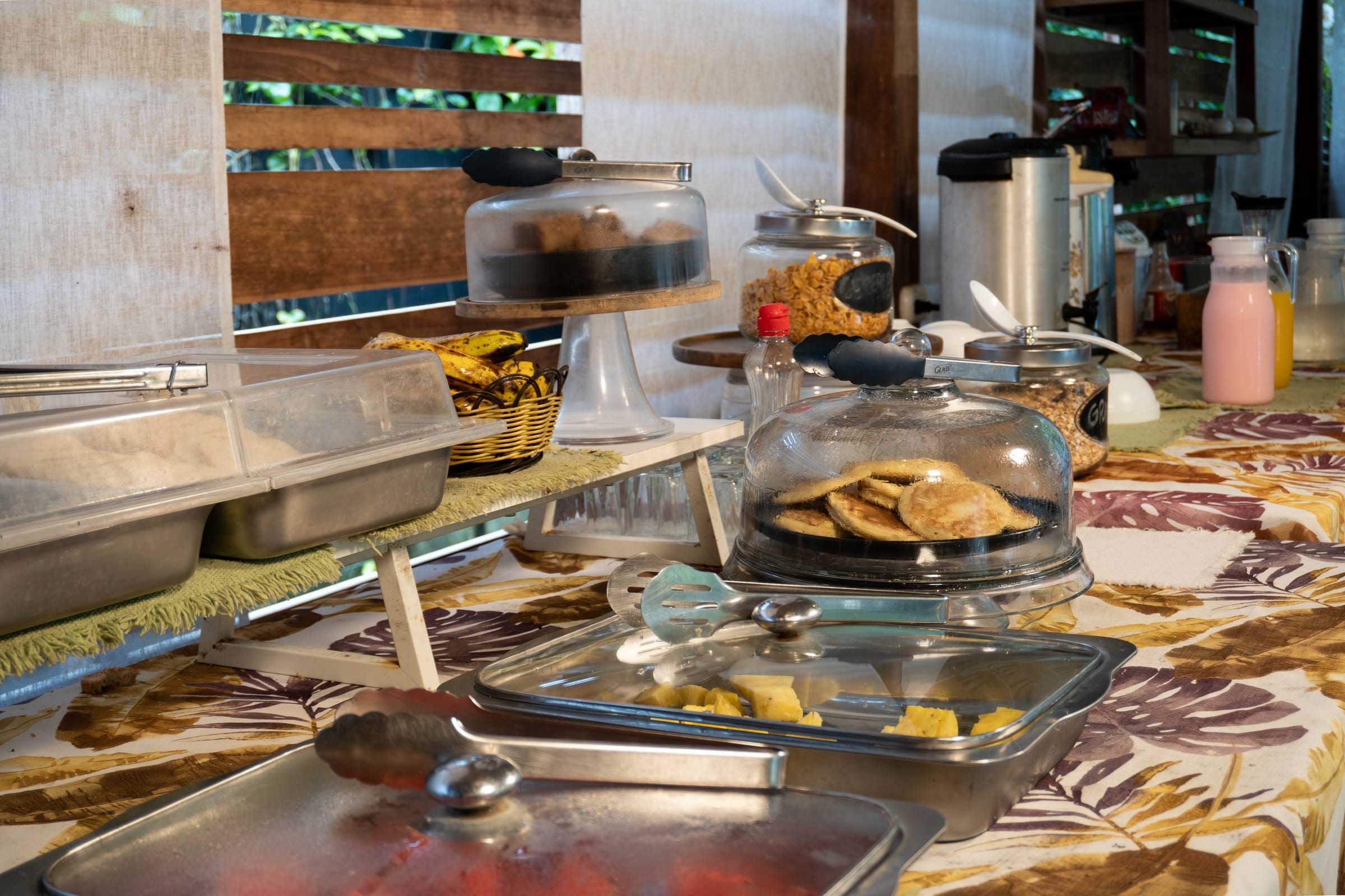 Breakfast buffet with covered pancake and pastry trays, fresh bananas, pineapple, cereal jars, and juice pitchers on a tropical-print tablecloth