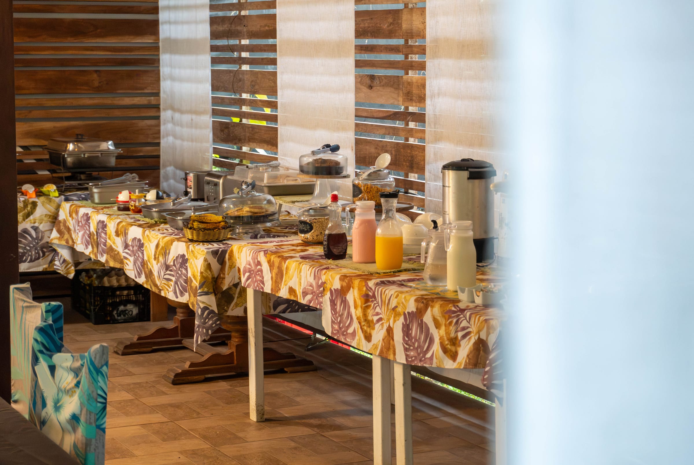 Long breakfast buffet table with chafing dishes, a coffee urn, fruit juices, and condiments set against a wood-slat and linen backdrop