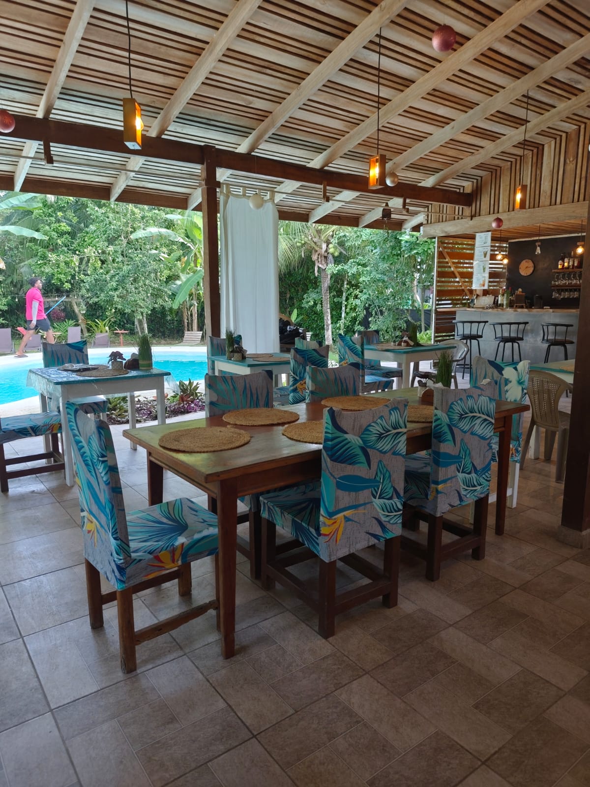 Covered open-air restaurant with wooden dining tables and tropical-print chairs, overlooking the pool and bar