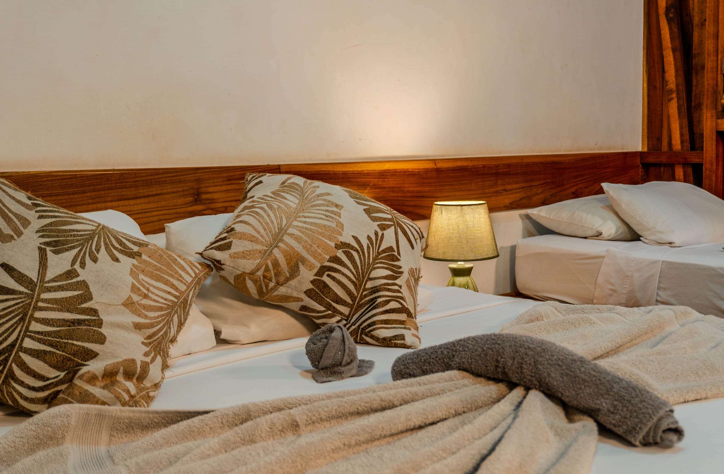 Lado Mar room bed with folded-towel decoration and leaf-embroidered pillows, a lit table lamp, and a second bed beside a wood-paneled wall
