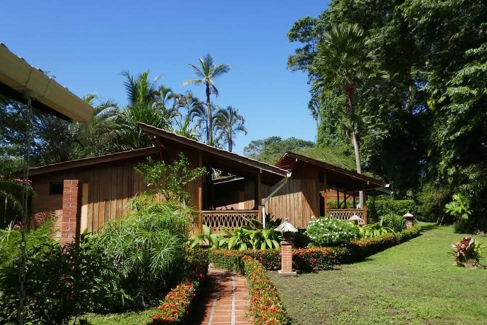 Wooden cabins with covered porches along a flower-lined brick path, palm trees and green lawn in the background