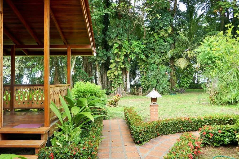 Wooden covered porch with a railing overlooking a brick garden path and a lawn dotted with tall palms
