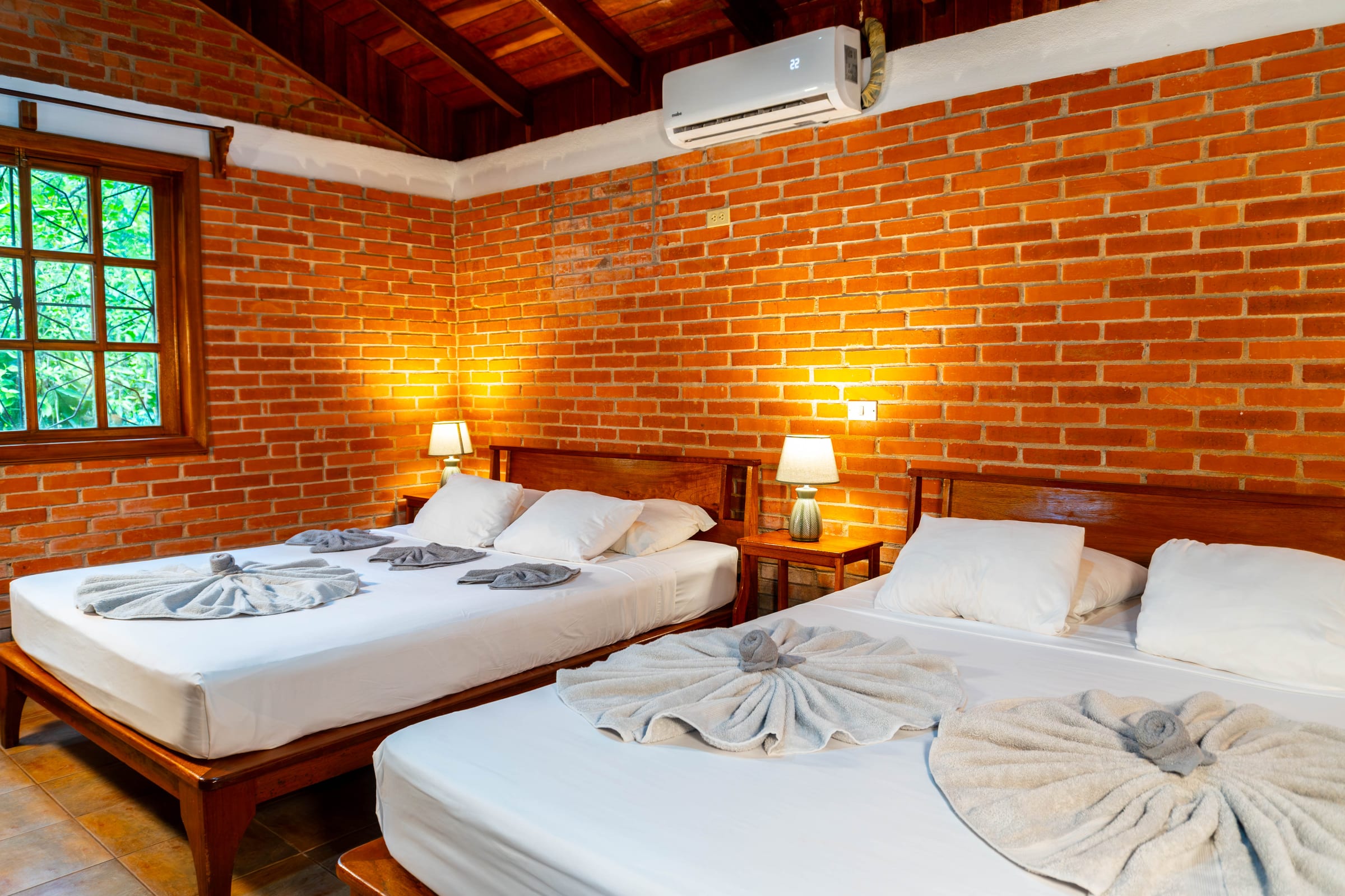 Lado Mar room interior with exposed brick walls, two beds dressed with towel swans, a wall-mounted air conditioner, and a window looking into the garden